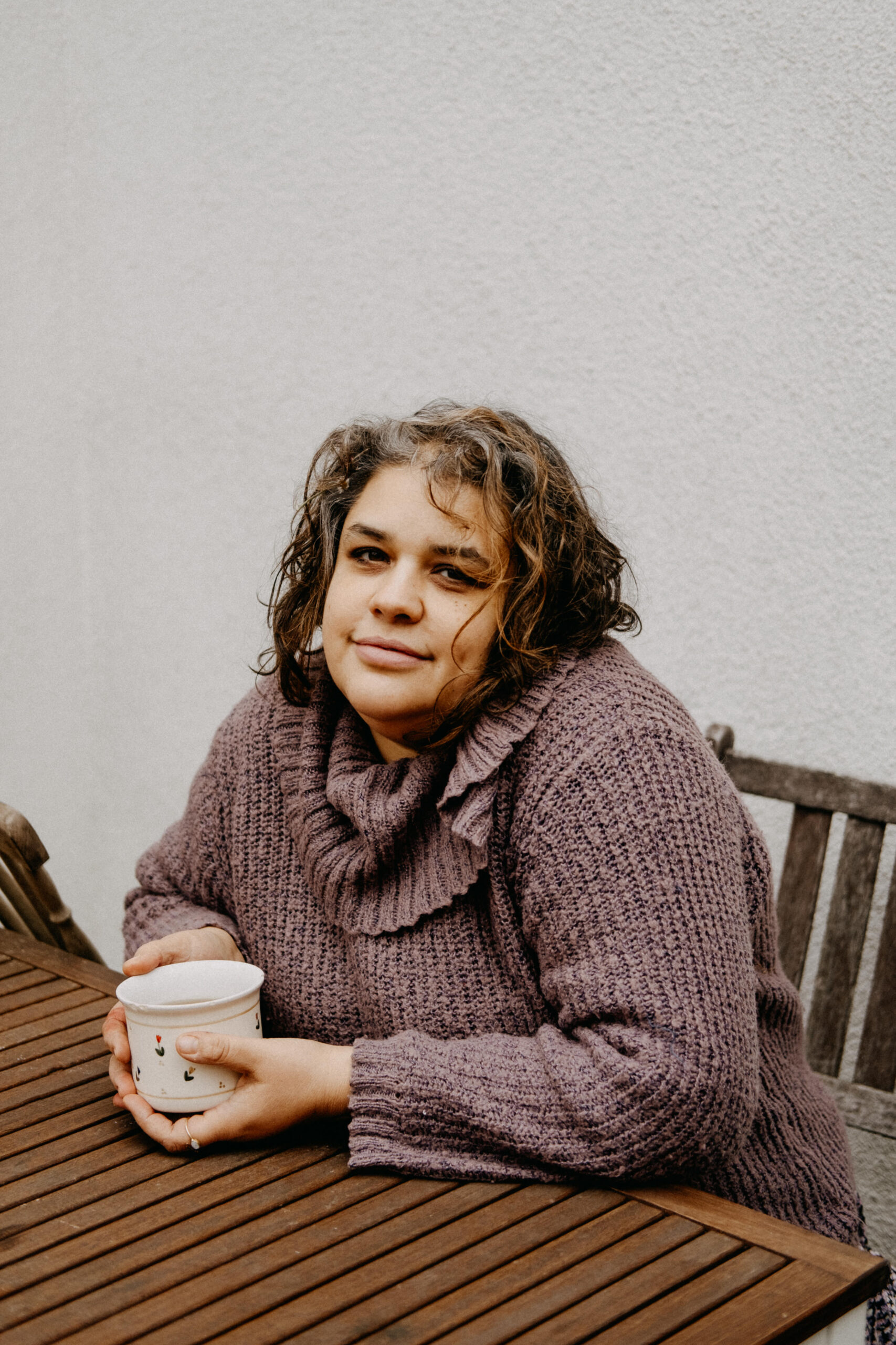 Ridina Ahmedova – portrait of a woman wearing a scarf and holding a mug at a wooden table.