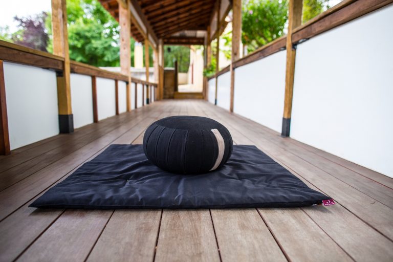 ZEN │ A Buddha sitting – meditation cushion on a dark mat in a covered wooden walkway.